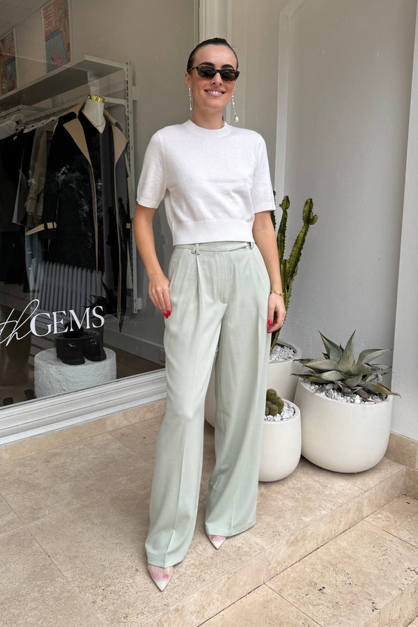 The image shows a smiling woman standing in front of a shop called "the GEMS." She is wearing a white short-sleeved crop top, light green wide-legged trousers, sunglasses, earrings, a bracelet, and pointed-toe heels. The scene includes potted cactus plants and a view into the shop window.