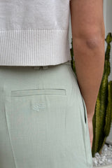 The image is a close-up shot of a person wearing a white cropped sweater and light green pants. The pants feature a back pocket with an embroidered logo. The setting seems to be indoors, with a cactus plant subtly visible in the background. The focus of the image is on the clothing and its details.
