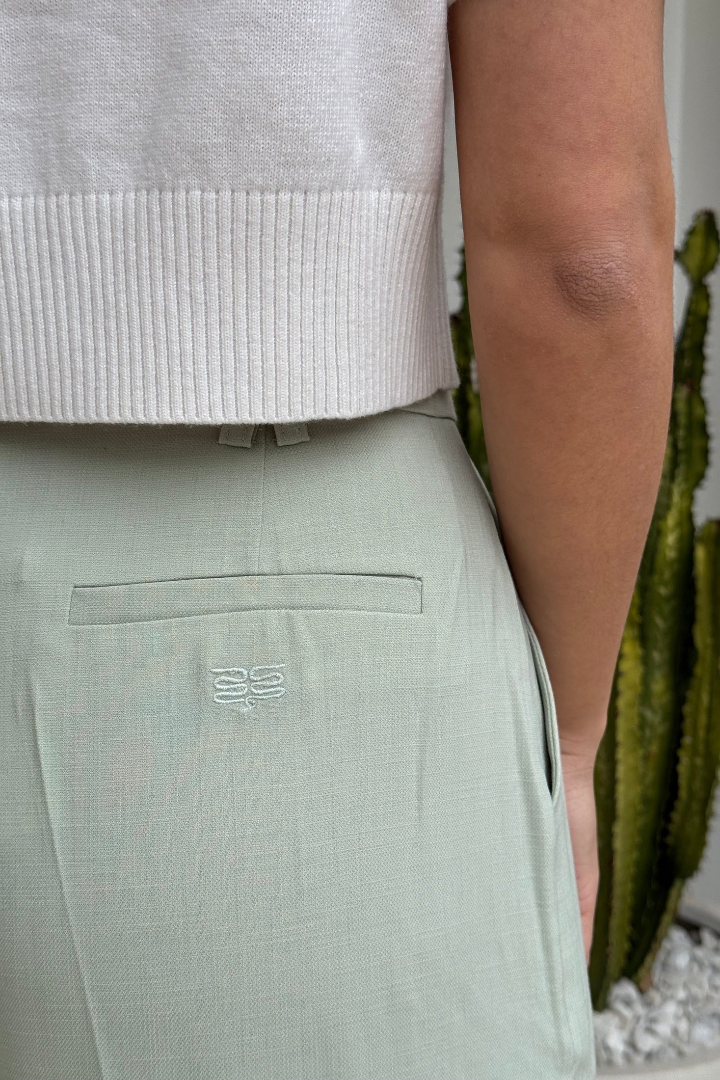 Woman in a light green coat and pants standing on a sidewalk with a building and hedge in the background