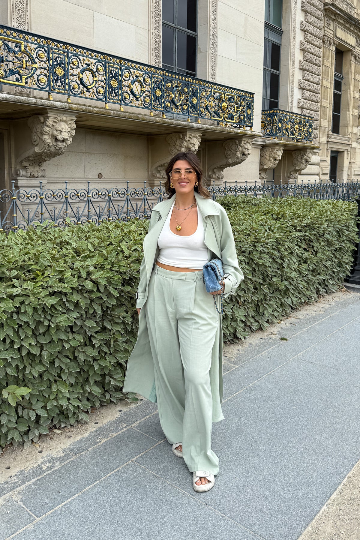 Woman in a light green coat and pants standing on a sidewalk with a building and hedge in the background