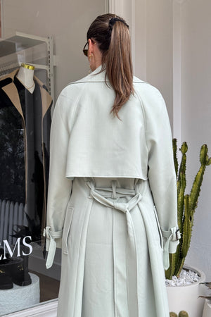 The image shows a woman from the back, wearing a light green trench coat outside of a retail store. Her brown hair is styled in a ponytail held by a black scrunchie, and she's wearing sunglasses and gold earrings. The backdrop includes a store window displaying merchandise, and a potted cactus. The letters "MS" are visible on the window, likely part of the store's branding.
