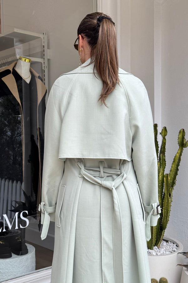 The image shows a woman from the back, wearing a light green trench coat outside of a retail store. Her brown hair is styled in a ponytail held by a black scrunchie, and she's wearing sunglasses and gold earrings. The backdrop includes a store window displaying merchandise, and a potted cactus. The letters "MS" are visible on the window, likely part of the store's branding.