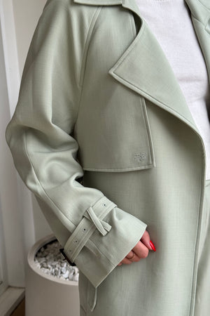 The image is a close-up shot of a light green trench coat worn over a white top. The trench coat features details like a cuff strap with a buckle, a pocket flap with a logo, and the person wearing the coat has red nail polish and a gold ring. The photo was likely taken indoors, with a plain background. The overall impression is one of understated elegance and attention to detail.