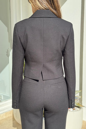 The image showcases a woman from the back, wearing a tailored black blazer and matching pants with buttoned cuffs on the blazer and an "EB" logo on the pants. The scene is set indoors against a white wall and window, with a potted succulent visible on the right, creating a minimalist backdrop that puts the focus on the clothing.