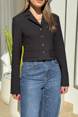 The image showcases a woman wearing a black blazer and blue jeans, accessorized with gold jewelry. She is standing near a white wall with a cactus plant in a pot visible in the frame. The image is well-lit, highlighting the colors of the clothing and accessories.