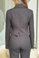 The image showcases a woman from the back, wearing a tailored black blazer and matching pants with buttoned cuffs on the blazer and an "EB" logo on the pants. The scene is set indoors against a white wall and window, with a potted succulent visible on the right, creating a minimalist backdrop that puts the focus on the clothing.