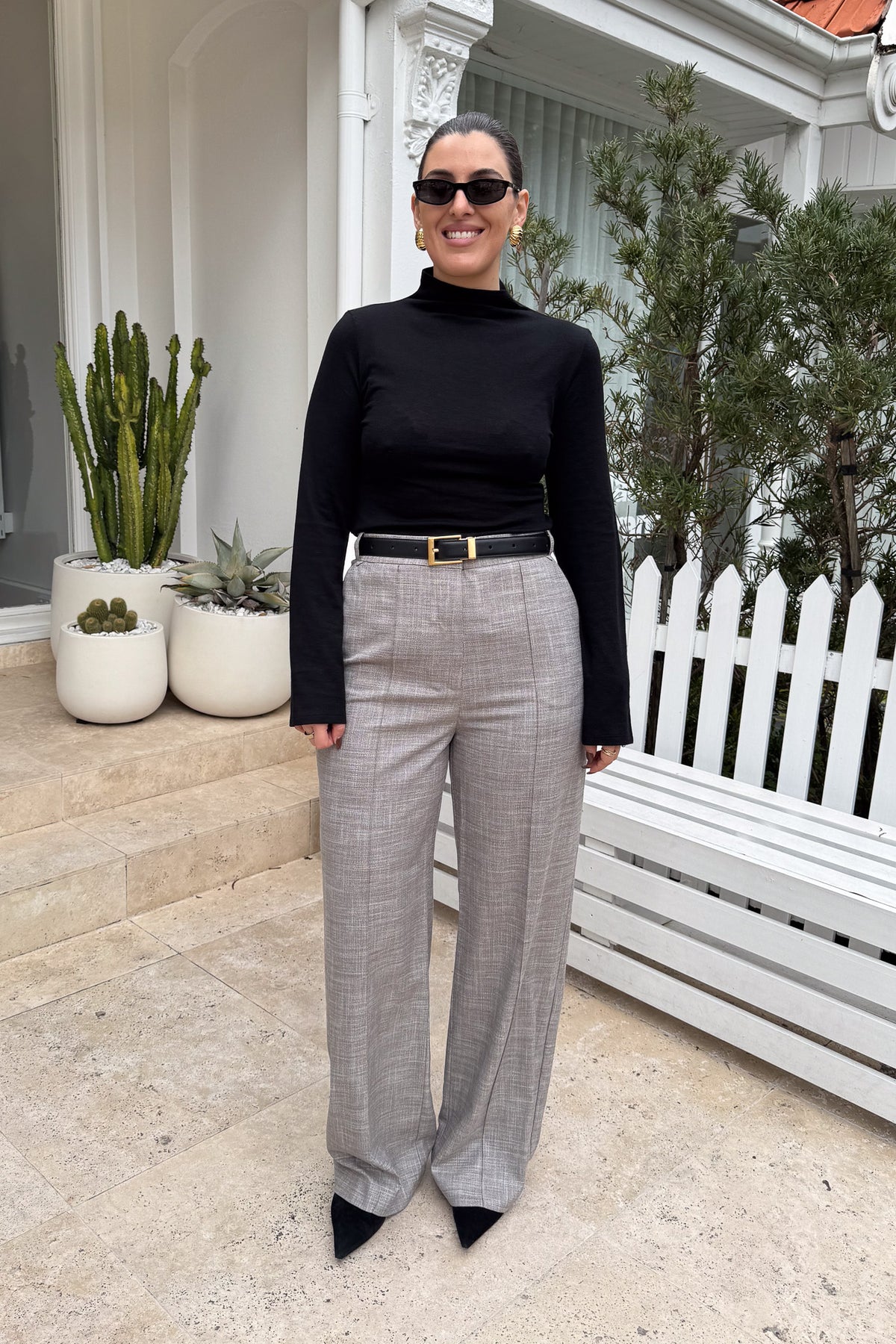 The image features a woman in a black turtleneck and grey pants with a black and gold belt, standing outdoors in front of a building. She is wearing sunglasses and gold earrings. The setting includes white plant pots containing cacti and succulents, along with a white picket fence and bench. The woman appears confident and stylish, with a neutral yet modern color scheme enhancing the overall aesthetic.