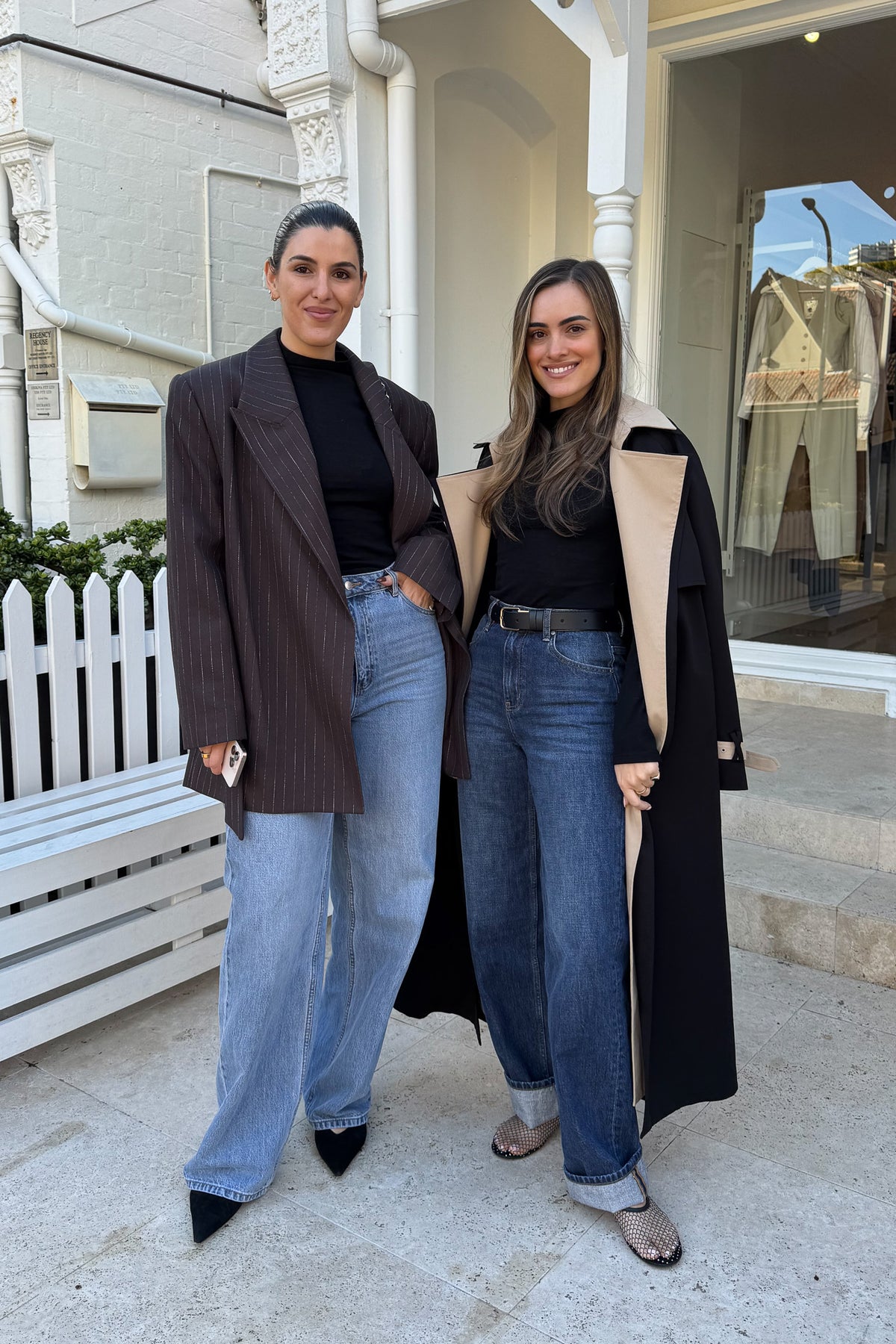 Two women are standing outdoors in front of a white building, likely a store called "Regency House". The woman on the left wears a brown pinstripe blazer, black turtleneck, and light blue jeans, holding a cell phone. The woman on the right wears a black trench coat with beige lining, a black turtleneck, dark blue jeans, and a black belt. Both women are smiling and appear relaxed.