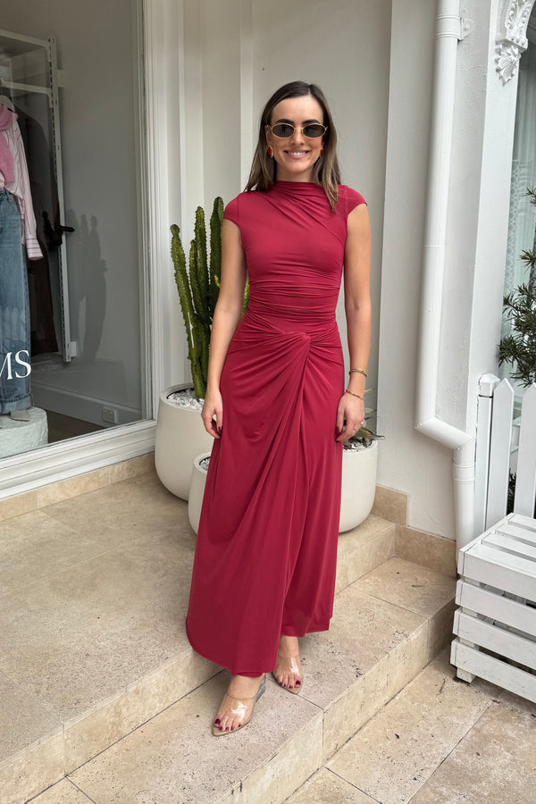 The image shows a smiling woman with brown hair wearing a red dress, sunglasses, and clear heels, posing on steps outside a building. The building appears to be a retail store with clothing visible through the window. A cactus in a white pot stands beside the steps. The dominant colors are red, white, and green.