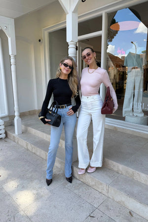 The Polly Long Sleeve top in pink. Woman in a pink top and white pants standing outside a store with plants and a bench in the background.
