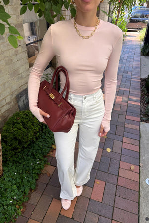 The Polly Long Sleeve top in pink. Woman in a pink top and white pants standing outside a store with plants and a bench in the background.