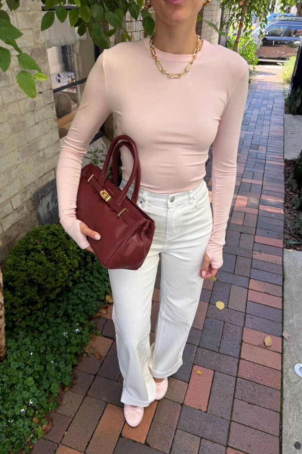 The Polly Long Sleeve top in pink. Woman in a pink top and white pants standing outside a store with plants and a bench in the background.