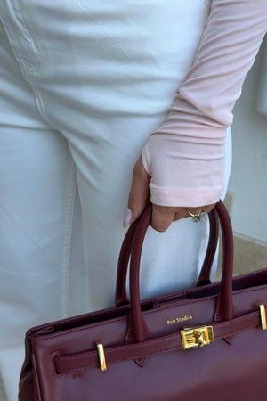 The Polly Long Sleeve top in pink. Woman in a pink top and white pants standing outside a store with plants and a bench in the background.