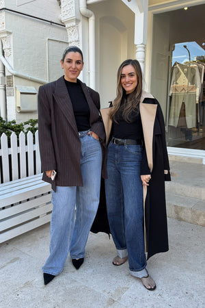 Two women are standing outdoors in front of a white building, likely a store called "Regency House". The woman on the left wears a brown pinstripe blazer, black turtleneck, and light blue jeans, holding a cell phone. The woman on the right wears a black trench coat with beige lining, a black turtleneck, dark blue jeans, and a black belt. Both women are smiling and appear relaxed.