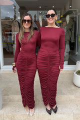 Two women pose in front of a store wearing matching maroon, long-sleeved, ruched dresses. The woman on the left has long light brown hair, brown sunglasses, a gold cuff, and clear high heels, while the woman on the right has her dark hair pulled back, dark sunglasses, and black mesh high heels. Both women are smiling, and the scene is set outside of a store with mannequins in the window.