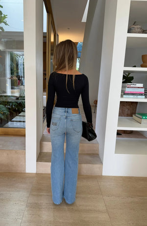 A woman stands in a bright, minimalist room wearing the Aria Boat Neck Long Sleeve Top Black with light-wash flared jeans.