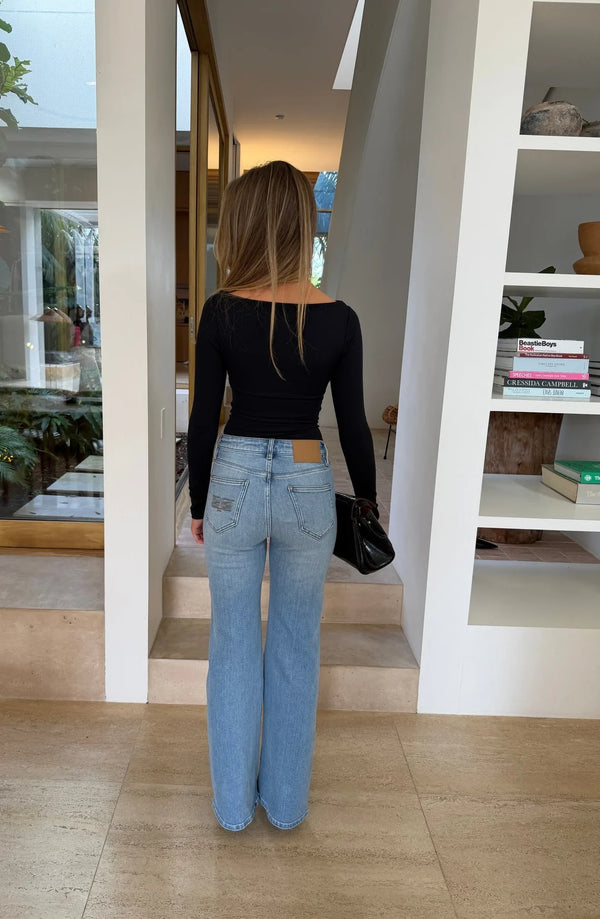 A woman stands in a bright, minimalist room wearing the Aria Boat Neck Long Sleeve Top Black with light-wash flared jeans.