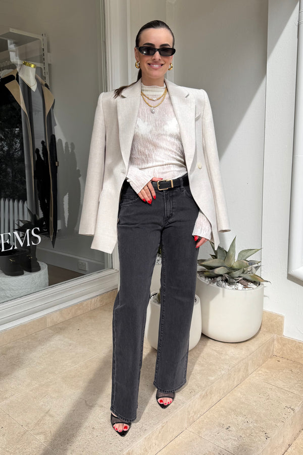 The image shows a stylish woman standing outside a building, likely a store, as indicated by the "EMS" text on the window. She is wearing a beige blazer, a light-colored top, dark jeans with a black belt, and black heeled shoes. Her accessories include sunglasses and gold jewelry, such as a necklace and hoop earrings. The overall color scheme is neutral, with beige, black, and white tones. The woman is smiling, projecting a confident and fashionable appearance.