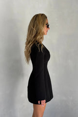 A woman in a fitted Bobbie Long Sleeve Mini Dress Black poses in profile wearing dark sunglasses against a textured gray wall.