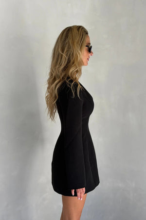 A woman in a fitted Bobbie Long Sleeve Mini Dress Black poses in profile wearing dark sunglasses against a textured gray wall.