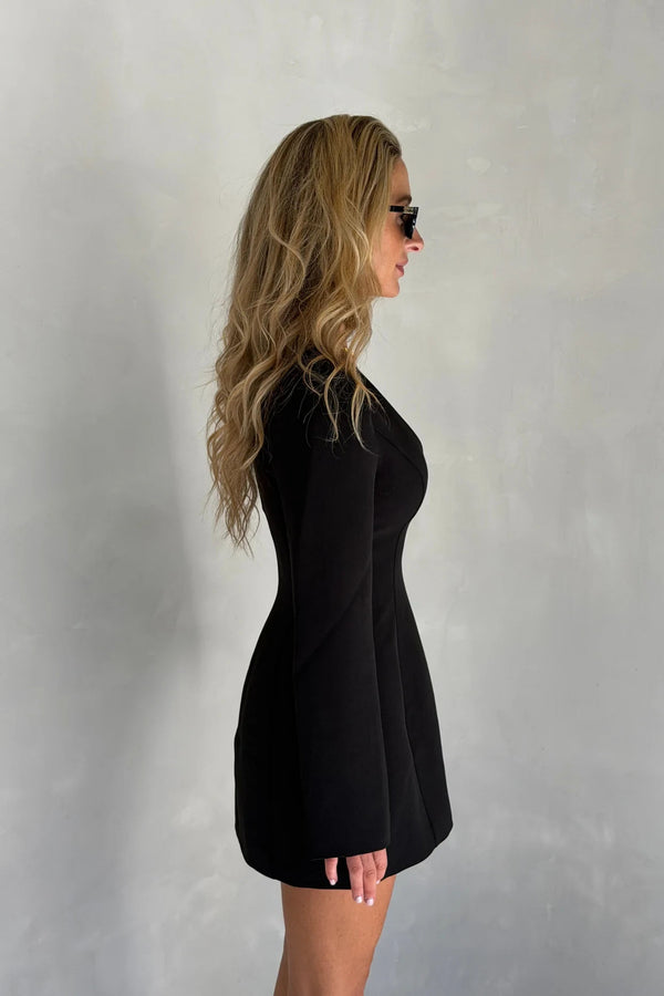 A woman in a fitted Bobbie Long Sleeve Mini Dress Black poses in profile wearing dark sunglasses against a textured gray wall.