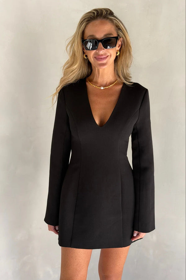 A woman wears a tailored black Bobbie Long Sleeve Mini Dress with a deep V-neck, gold jewelry, and Gucci sunglasses against a grey backdrop.