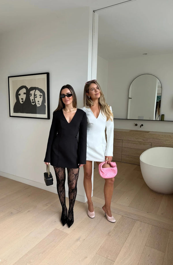 Two women pose in a modern home, one wearing the black Bobbie Long Sleeve Mini Dress with lace tights and heels.