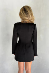 Woman with long blonde hair wearing a black Bobbie long sleeve mini dress with sharp shoulders against a light grey background.