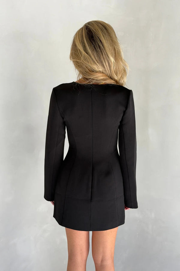 Woman with long blonde hair wearing a black Bobbie long sleeve mini dress with sharp shoulders against a light grey background.