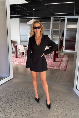Woman wearing a black Bobbie long sleeve mini dress with pointed heels and sunglasses in a modern office setting.