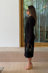 A woman in a fitted black Carissa Tailored Midi Dress stands in a minimalist room holding a quilted Chanel bag with gold accents.