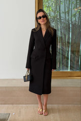 A woman wears the Carissa Tailored Midi Dress Black with gold heels and a quilted black handbag, standing by a glass partition with bamboo.