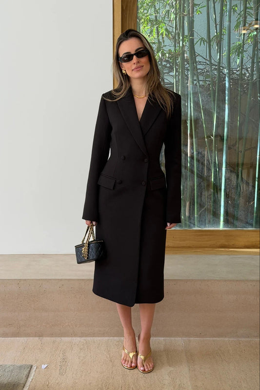 A woman wears the Carissa Tailored Midi Dress Black with gold heels and a quilted black handbag, standing by a glass partition with bamboo.