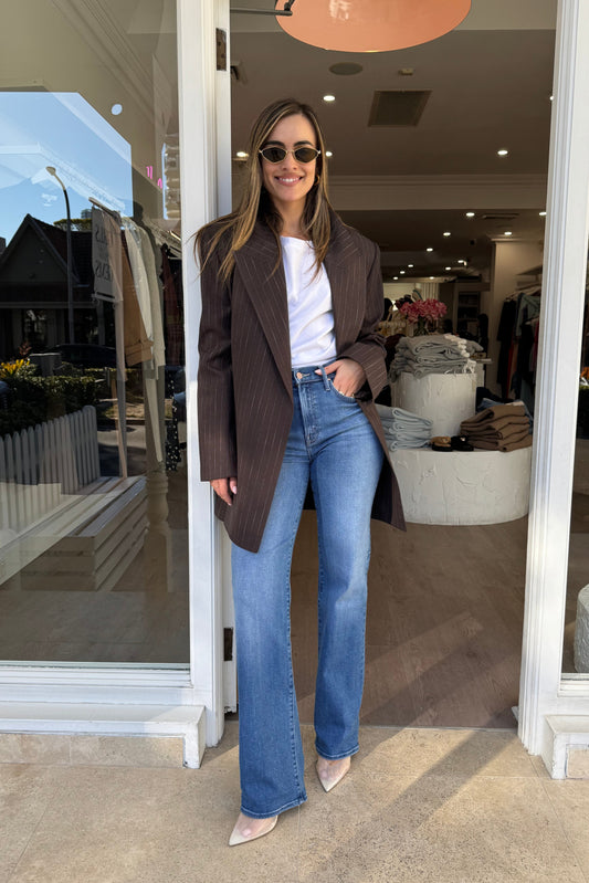 The image captures a smiling woman with long brown hair posing confidently in the doorway of a clothing store. She is wearing a brown pinstripe blazer, blue jeans, a white t-shirt, sunglasses, and light-colored heeled shoes. The store has a white-framed entrance, and the interior reveals clothing racks and mannequins. The scene is set on a light tan sidewalk, with reflections of a residential neighborhood visible in the store windows. The overall impression is stylish and inviting.