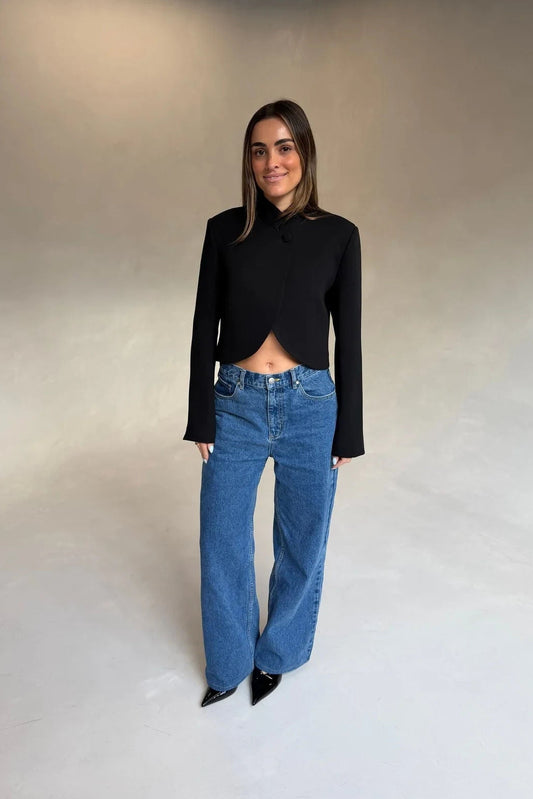 A woman wears the Charlotte Cropped Blazer Top Black with high-waisted blue jeans and black patent heels against a grey backdrop.