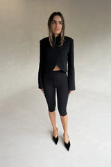 A woman wears a black Charlotte Cropped Blazer Top with a curved hem, paired with matching cycling shorts and patent leather pumps.