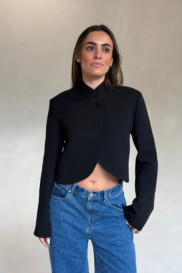 A young woman wears a black Charlotte Cropped Blazer Top with a high neck and single button, paired with blue denim jeans.