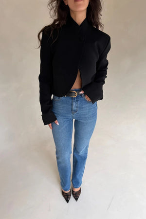 A woman wears a structured black Charlotte Cropped Blazer Top with medium-wash jeans, a black belt, and black mesh heels.