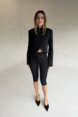 A woman wears a black Charlotte Cropped Blazer Top with capri leggings and patent leather heels against a grey background.