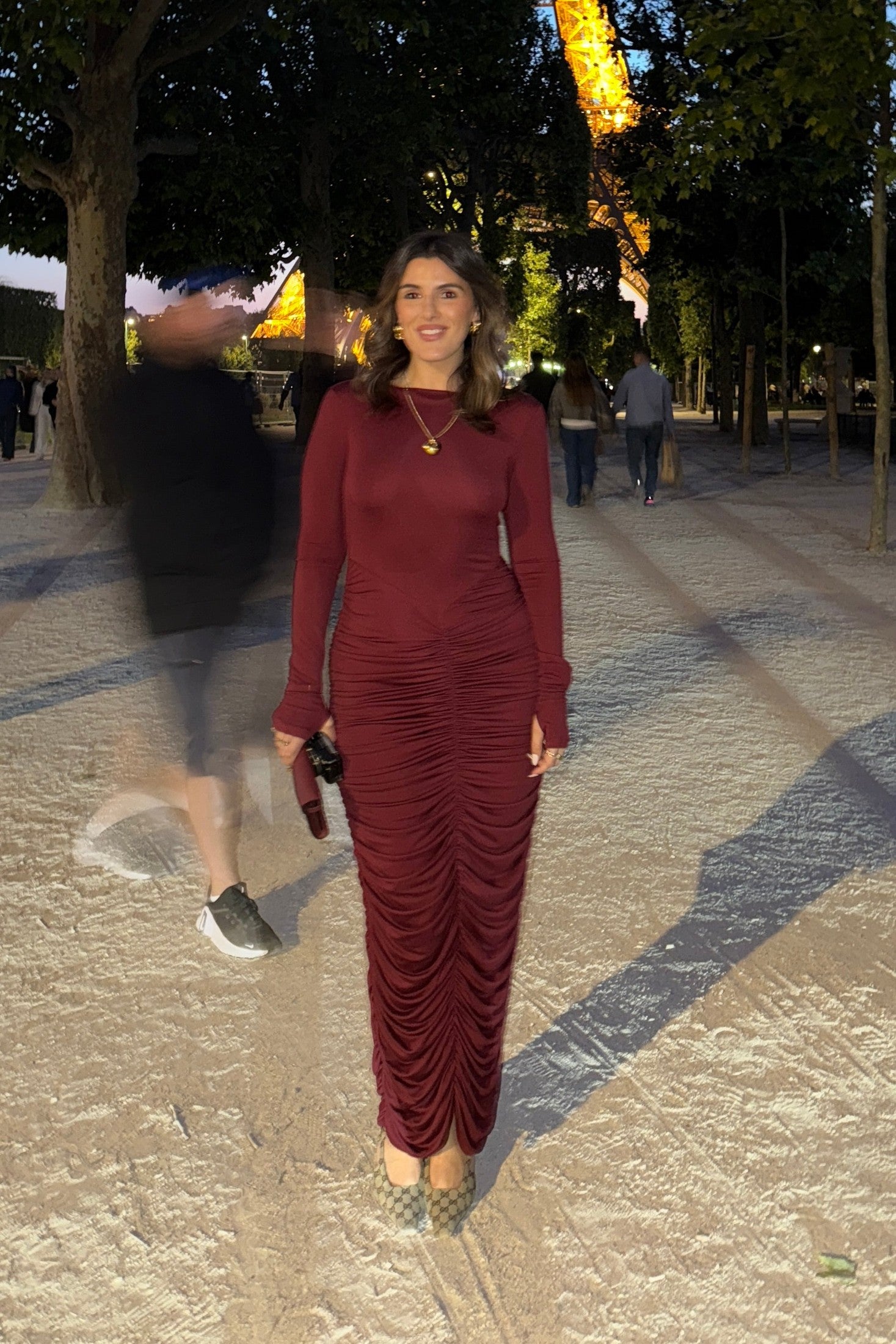 The image is a high-angle shot of a woman showcasing her outfit, which includes a burgundy, long-sleeved dress, a gold heart pendant necklace, a burgundy clutch, and Gucci high heels. The setting appears to be outdoors in front of a dark door. The colors are predominantly burgundy, gold, white, light brown, and gray.