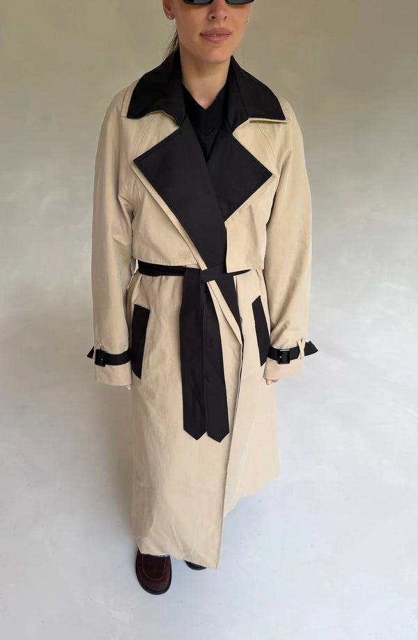 Beige Dakota trench coat with black contrast detailing, featuring a belted waist and classic collar.