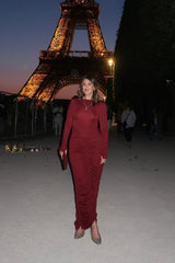 The image captures a woman in Paris posing in front of the Eiffel Tower during dusk. She is wearing a red long-sleeved dress and distinctive Gucci shoes, holding a clutch, and accessorized with a gold heart necklace. In the background, the illuminated Eiffel Tower stands tall, with other people and souvenir stands visible, giving context to the public tourist location.