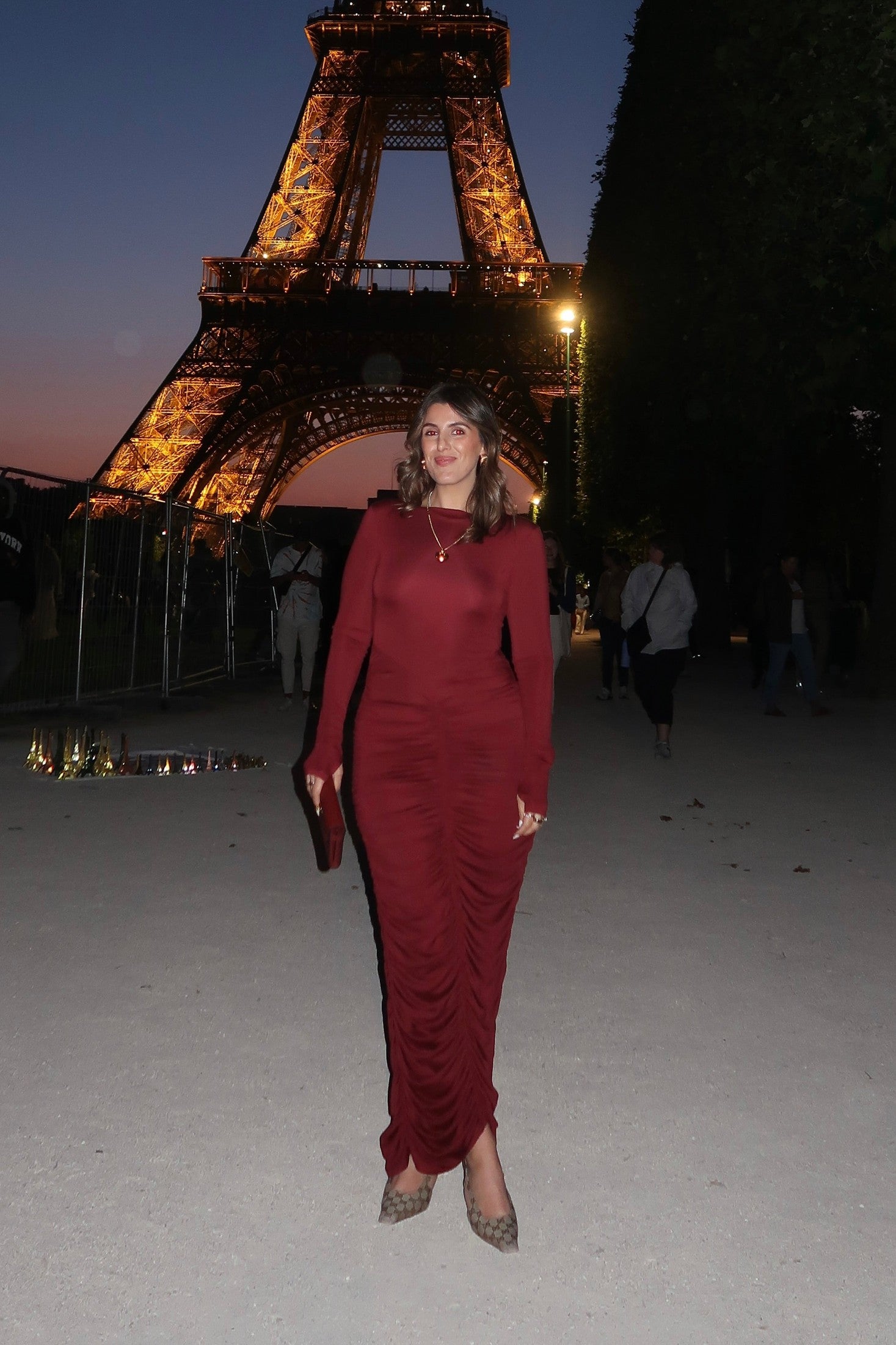 The image is a high-angle shot of a woman showcasing her outfit, which includes a burgundy, long-sleeved dress, a gold heart pendant necklace, a burgundy clutch, and Gucci high heels. The setting appears to be outdoors in front of a dark door. The colors are predominantly burgundy, gold, white, light brown, and gray.