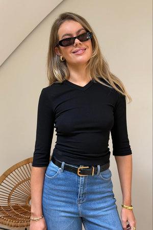 A blonde woman wears the Esme High V 3/4 Sleeve Top Black with blue jeans, gold jewelry, and Gucci sunglasses in a bright, minimalist room.