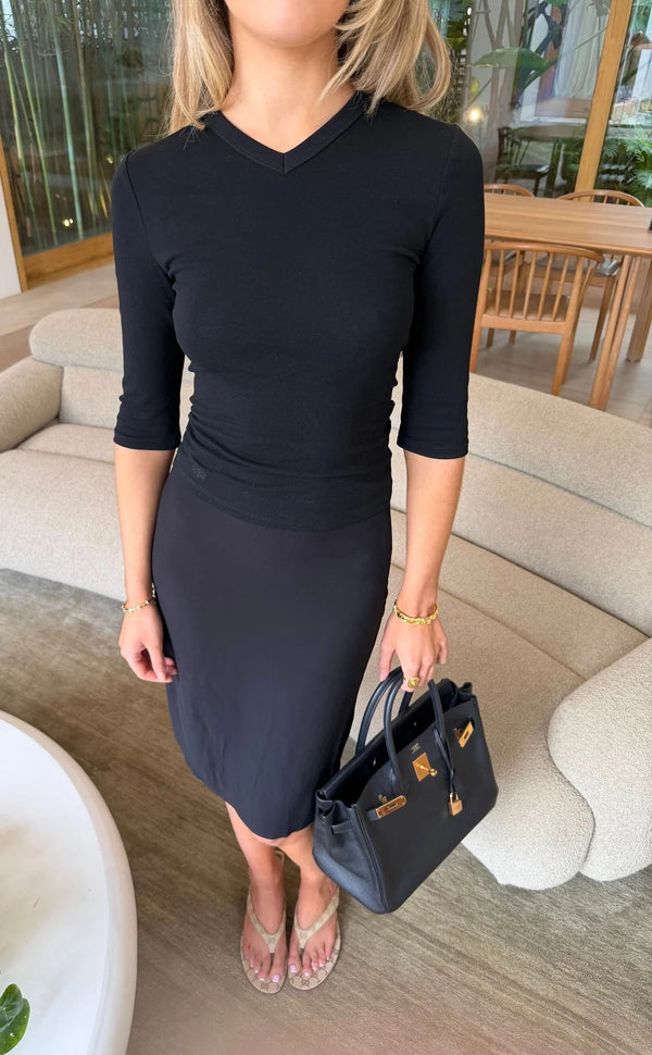 A woman wears the Esme High V 3/4 Sleeve Top Black with a black midi skirt and beige sandals in a minimalist living room.