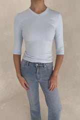A woman wears a light blue ribbed Esme High V 3/4 Sleeve Top paired with medium-wash jeans and white square-toe sandals.