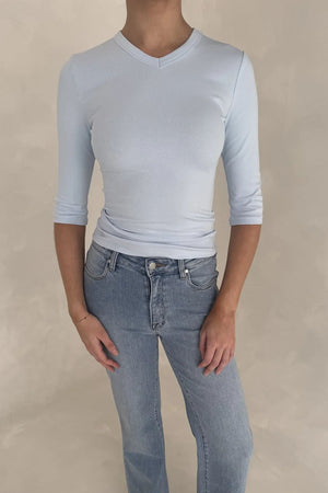 A woman wears a light blue ribbed Esme High V 3/4 Sleeve Top paired with medium-wash jeans and white square-toe sandals.