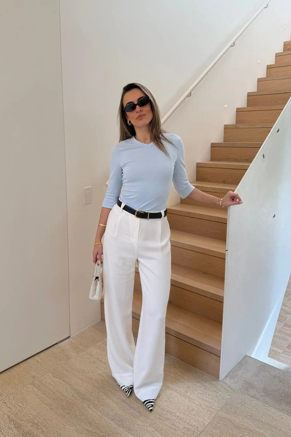 A woman stands on a modern staircase wearing the Esme High V light blue 3/4 sleeve top with white wide-leg trousers and zebra-print heels.