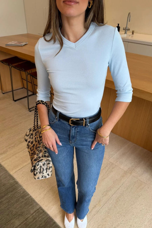 A woman wears the Esme High V 3/4 Sleeve Top in light blue paired with high-rise jeans, gold jewelry, and white pointed-toe heels.
