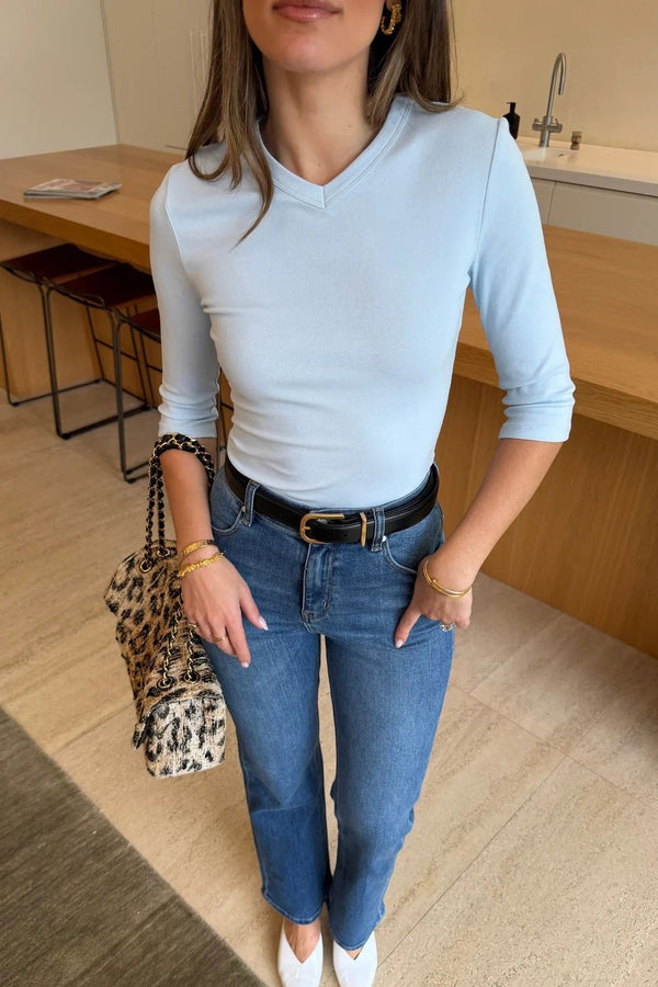 A woman wears the Esme High V 3/4 Sleeve Top in light blue paired with high-rise jeans, gold jewelry, and white pointed-toe heels.